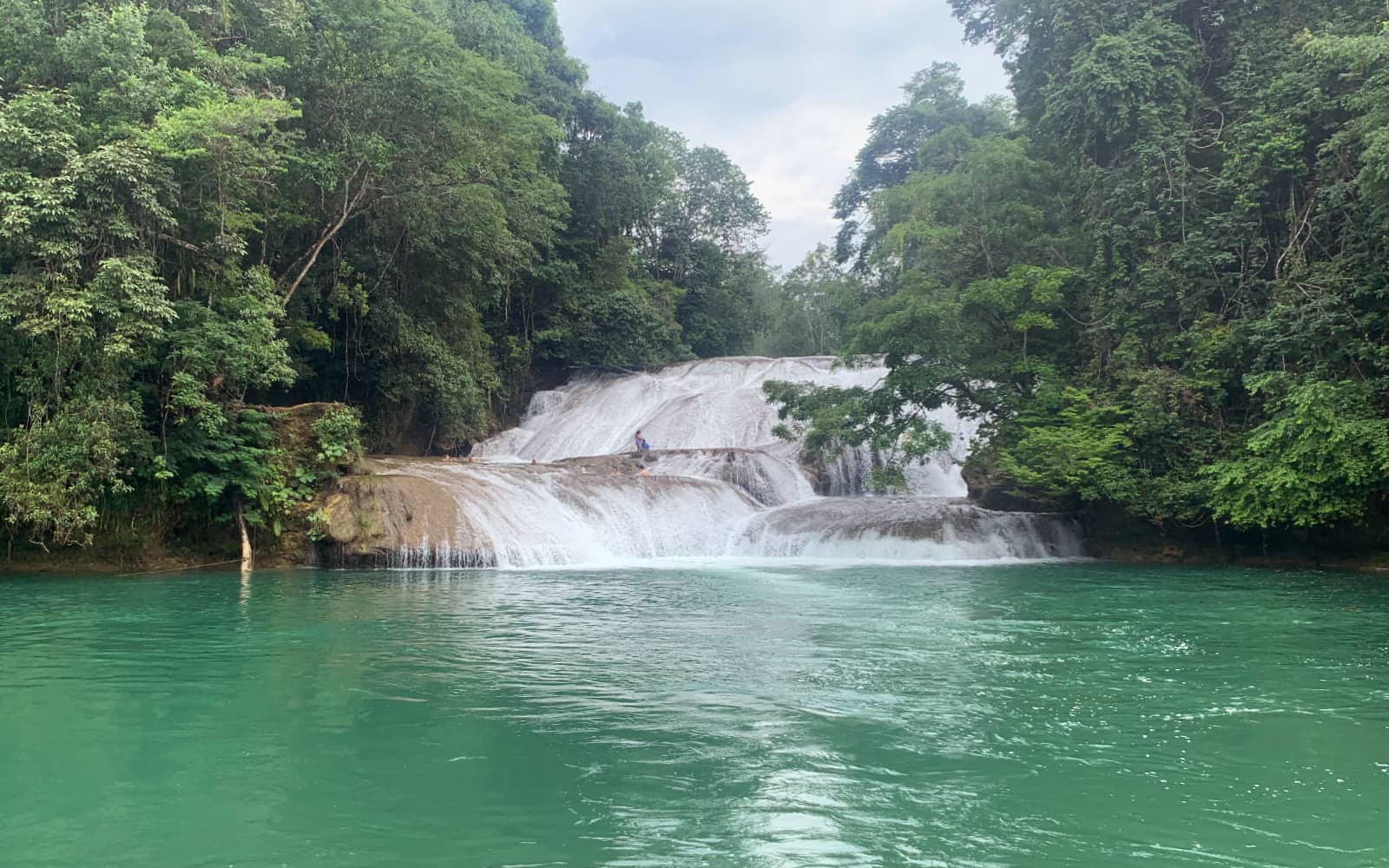 Large volume of water cascading over rocks into large turquoise pool with green trees on left and right side of water