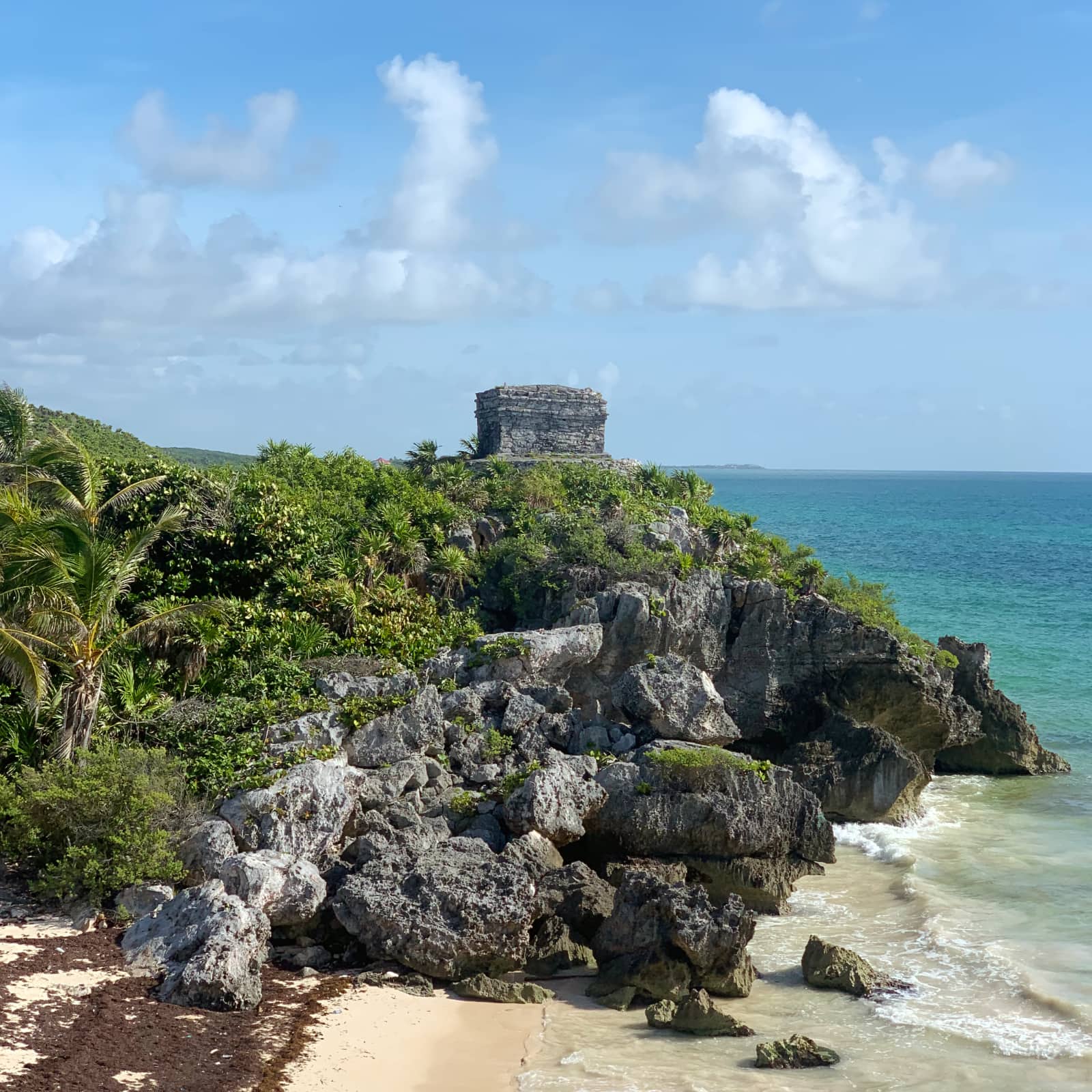 Small stone building perched on rocky cliff with palm trees in foreground and ocean in background