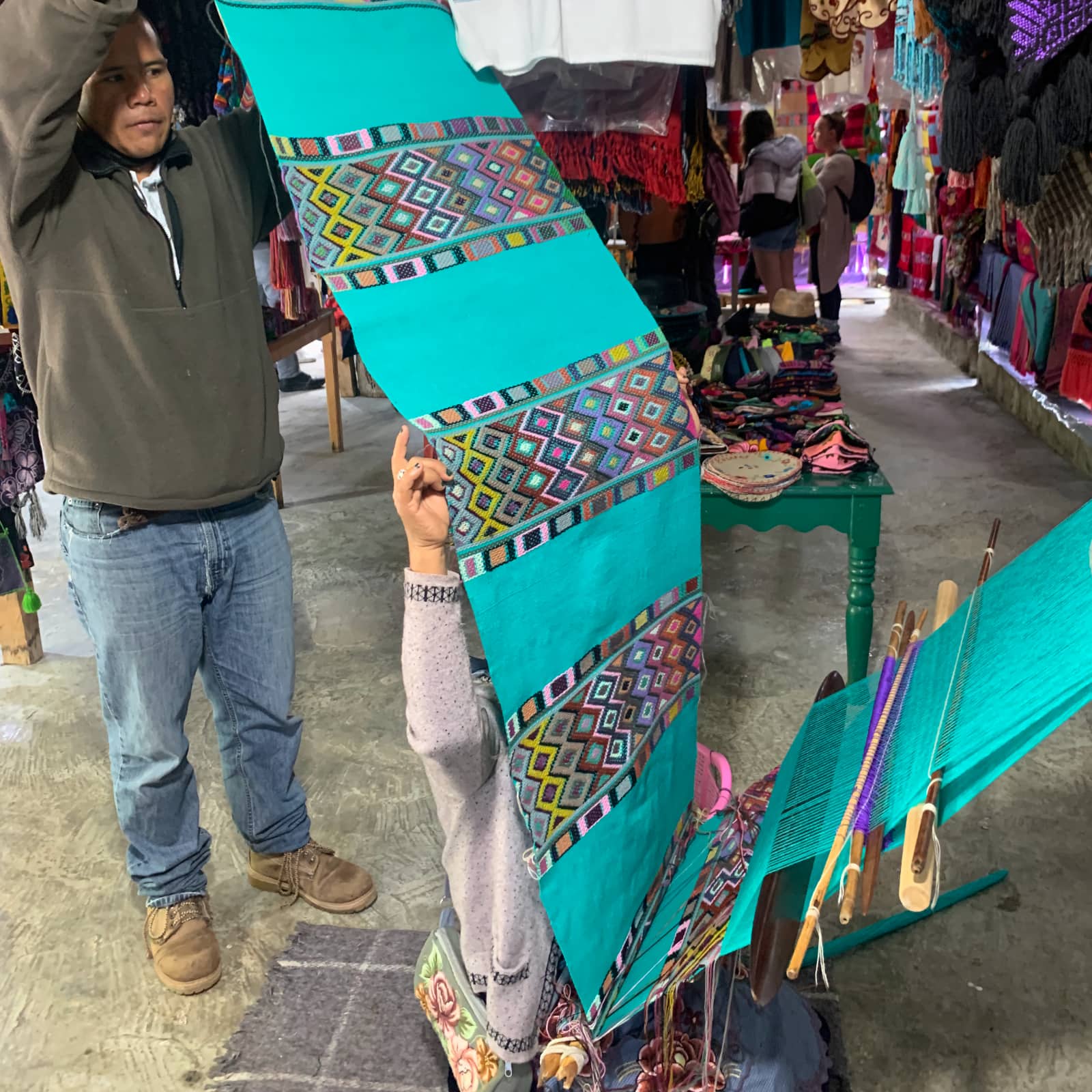 Long piece of textile being hand woven with a mix of turquoise, red, blue, and brown colours