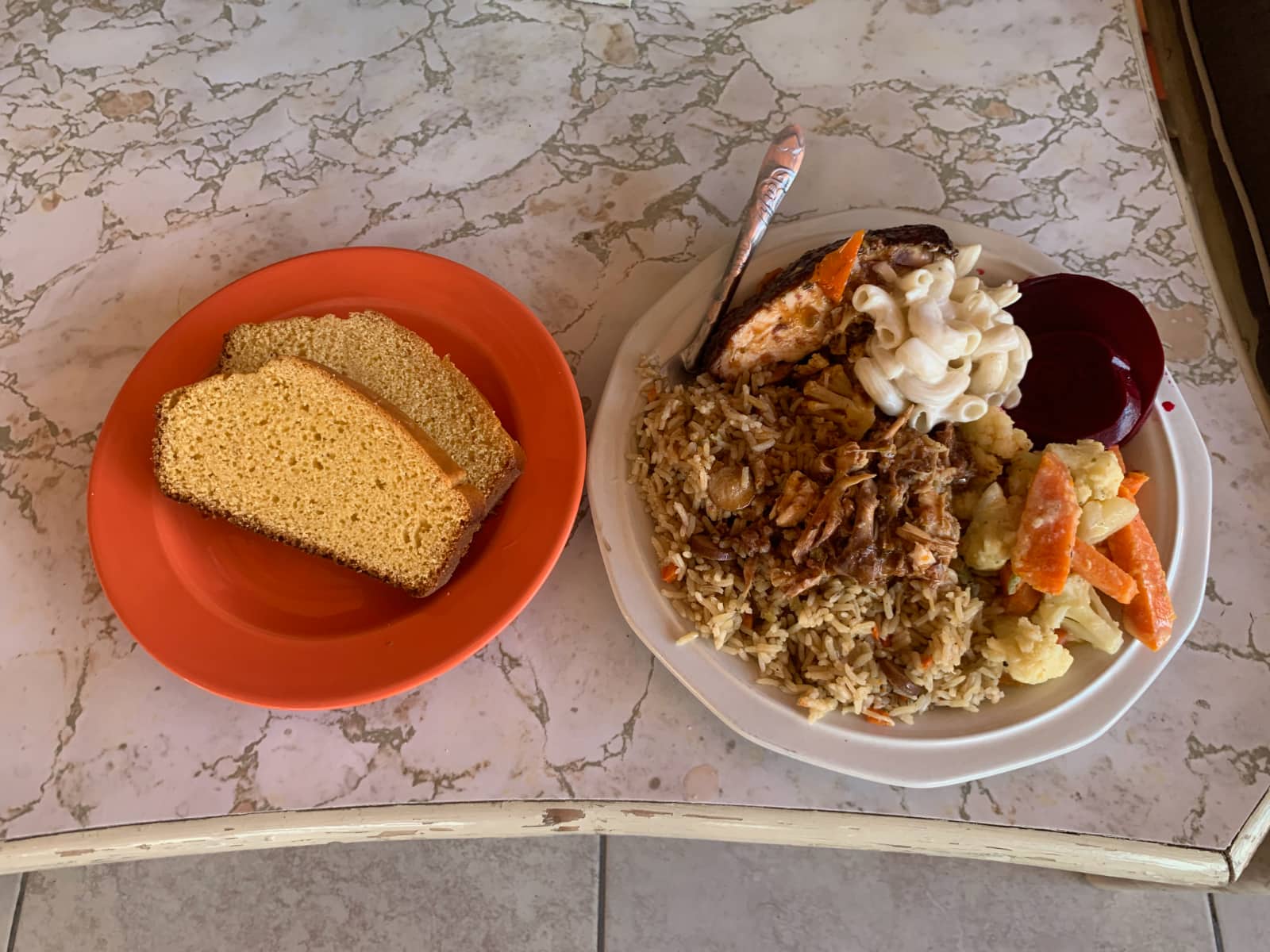 A red coloured plate containing two slices of corn bread and a white coloured plate containing rice, chicken, pasta salad, carrots and cauliflower, and roast pork.